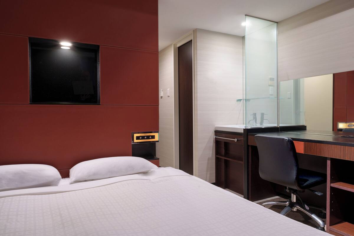 A hotel room with a bed, a desk with a chair, a mirror, and a small window.