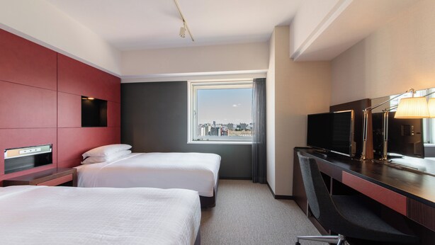 Habitación de hotel con dos camas, un escritorio con televisor, una ventana con vista a la ciudad y una pared de acento roja.