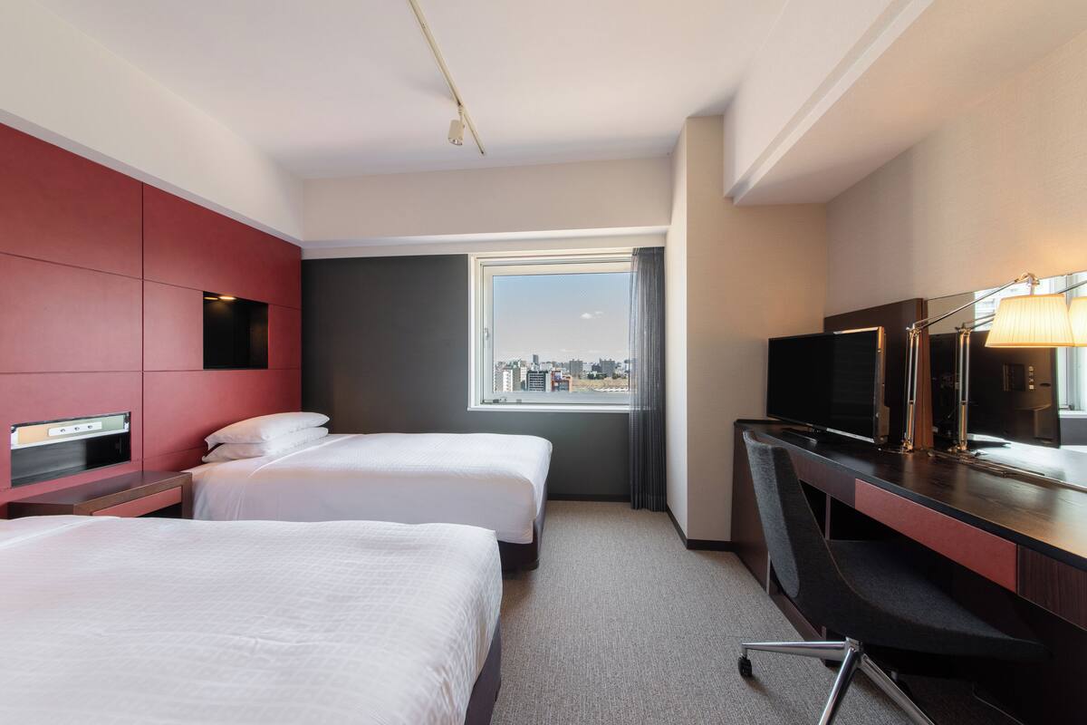A hotel room with two beds, a desk with a TV, a window with a city view, and a red accent wall.