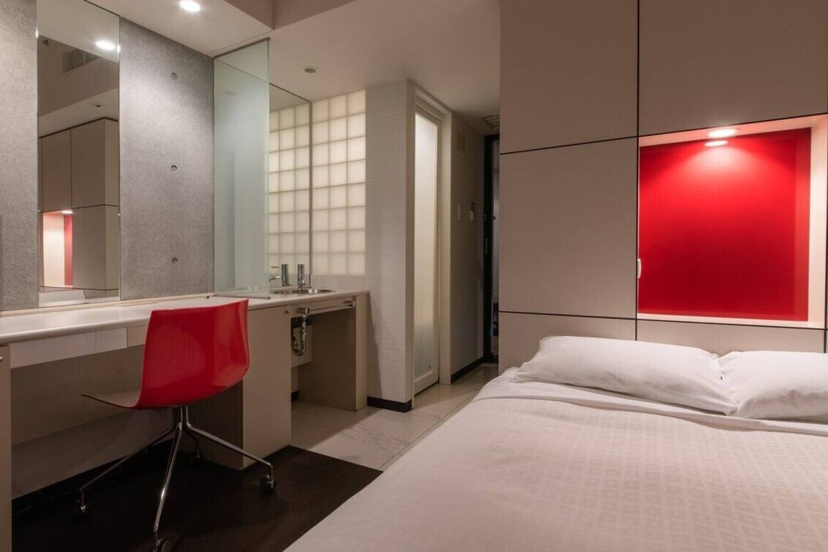 A modern hotel room with a bed, a desk with a chair, a large mirror, and a red accent wall.