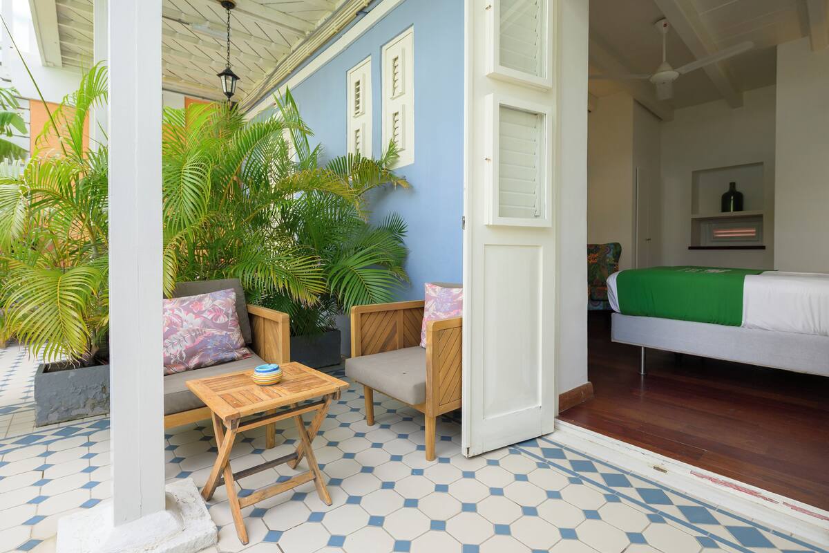 A room with a bed, a wooden table, and chairs, a potted plant, and a view of the outside.