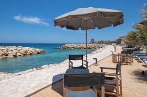 Un área de estar frente a la playa con una mesa y sillas bajo una sombrilla, con vista al mar y a la costa rocosa.