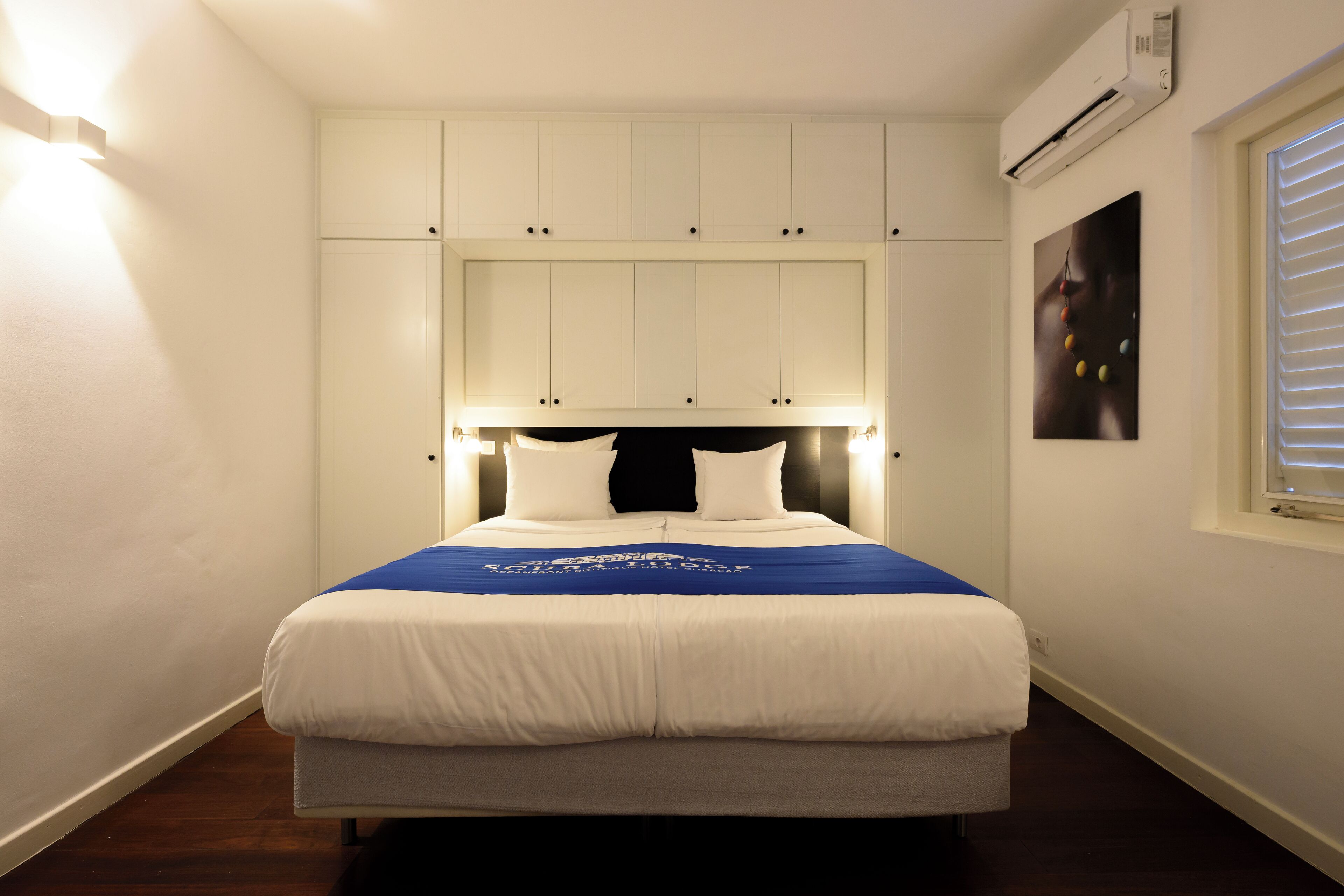 Een netjes opgemaakt bed met een blauw-witte sprei, een hoofdbord met ingebouwde verlichting en een raam met jaloezieën aan de rechterkant.