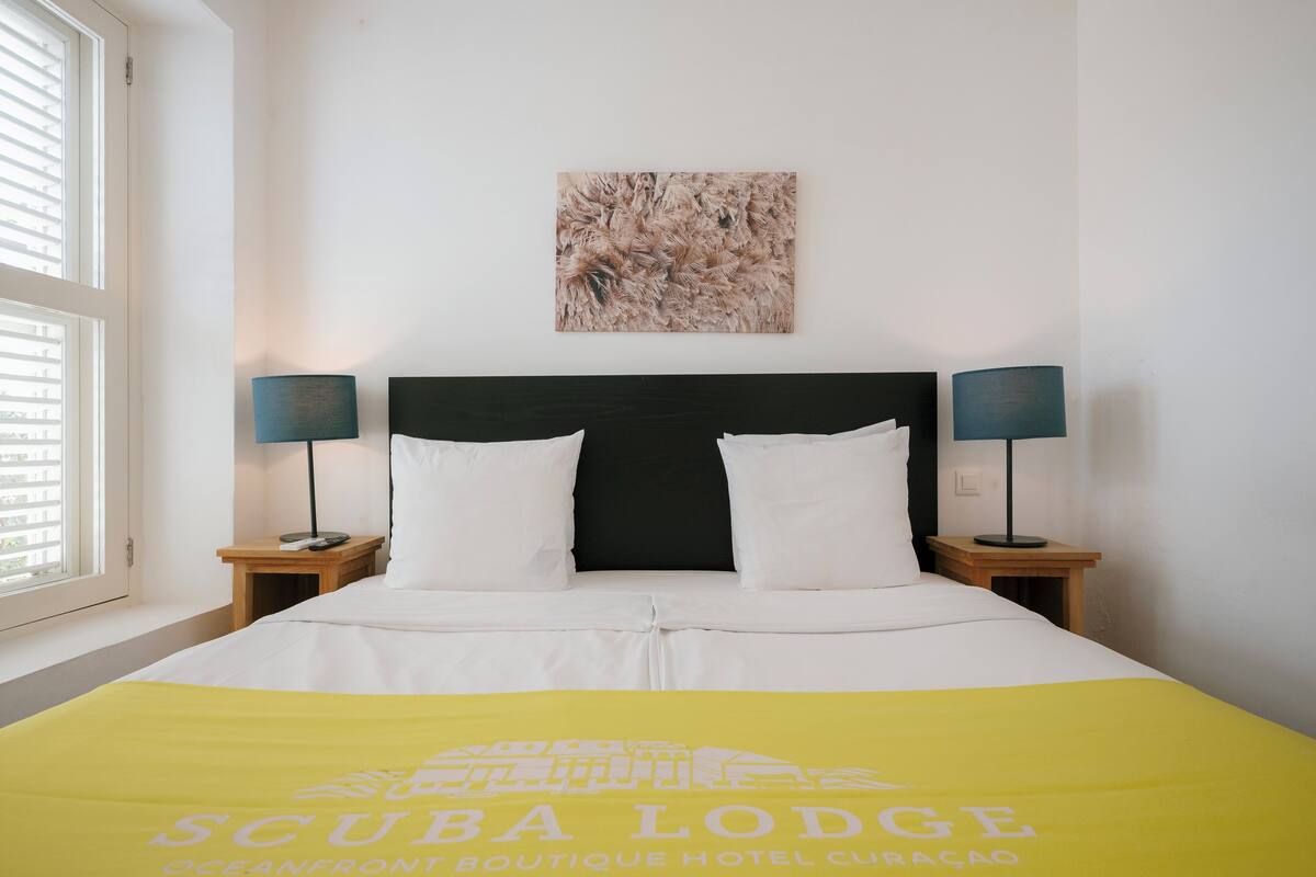 A hotel room with a bed, two bedside tables, a nightstand, a window with blinds, and a wall-mounted artwork.