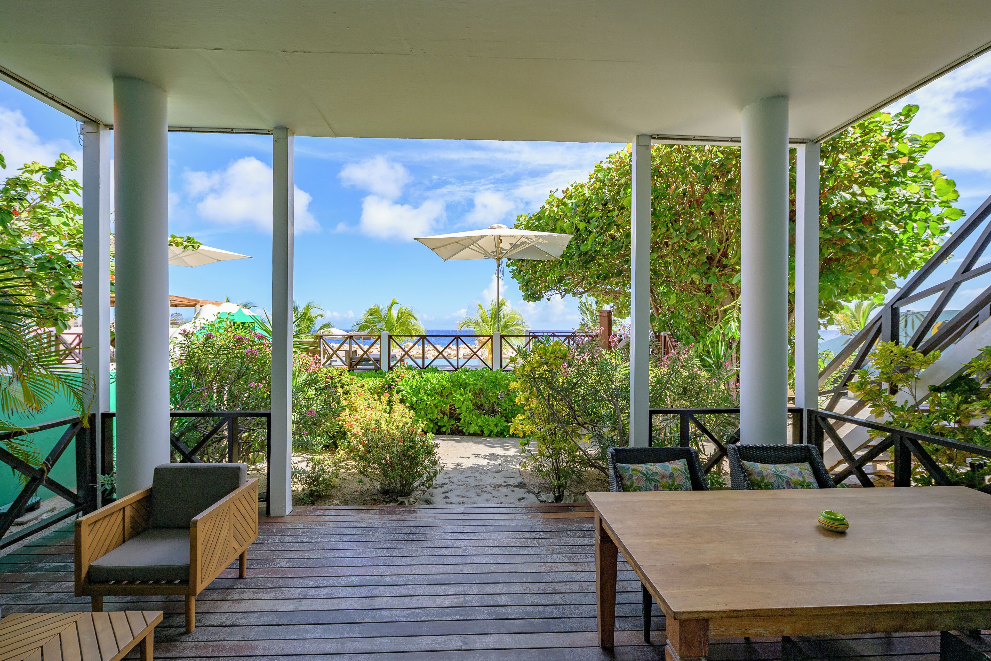 Eine überdachte Terrasse mit Holztablett und Stühlen, ein Weg zum Strand sowie Sonnenschirme, die Schatten spenden.