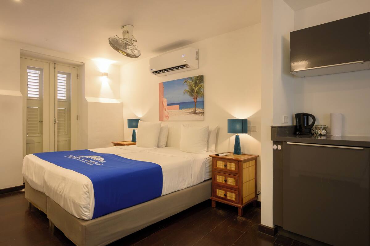 A hotel room with a bed, bedside tables, a wall-mounted air conditioner, a small kitchen area with a refrigerator, and a door with a window.