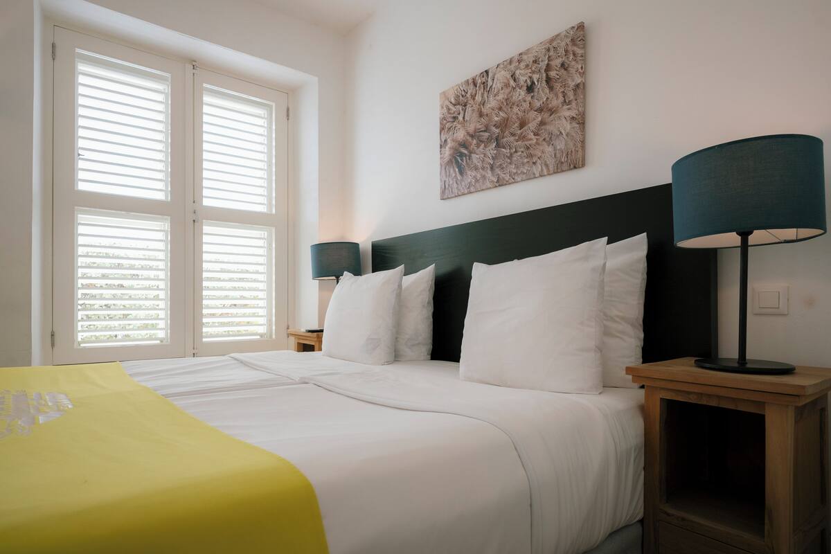 A neatly made bed with white linens, a green headboard, and a yellow throw blanket. There is a bedside table with a lamp and a wall-mounted artwork above the bed.