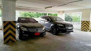 A covered parking area with two cars parked, concrete pillars with chevron patterns, and a view of greenery outside. - Poonchock Mansion (Bangkok)