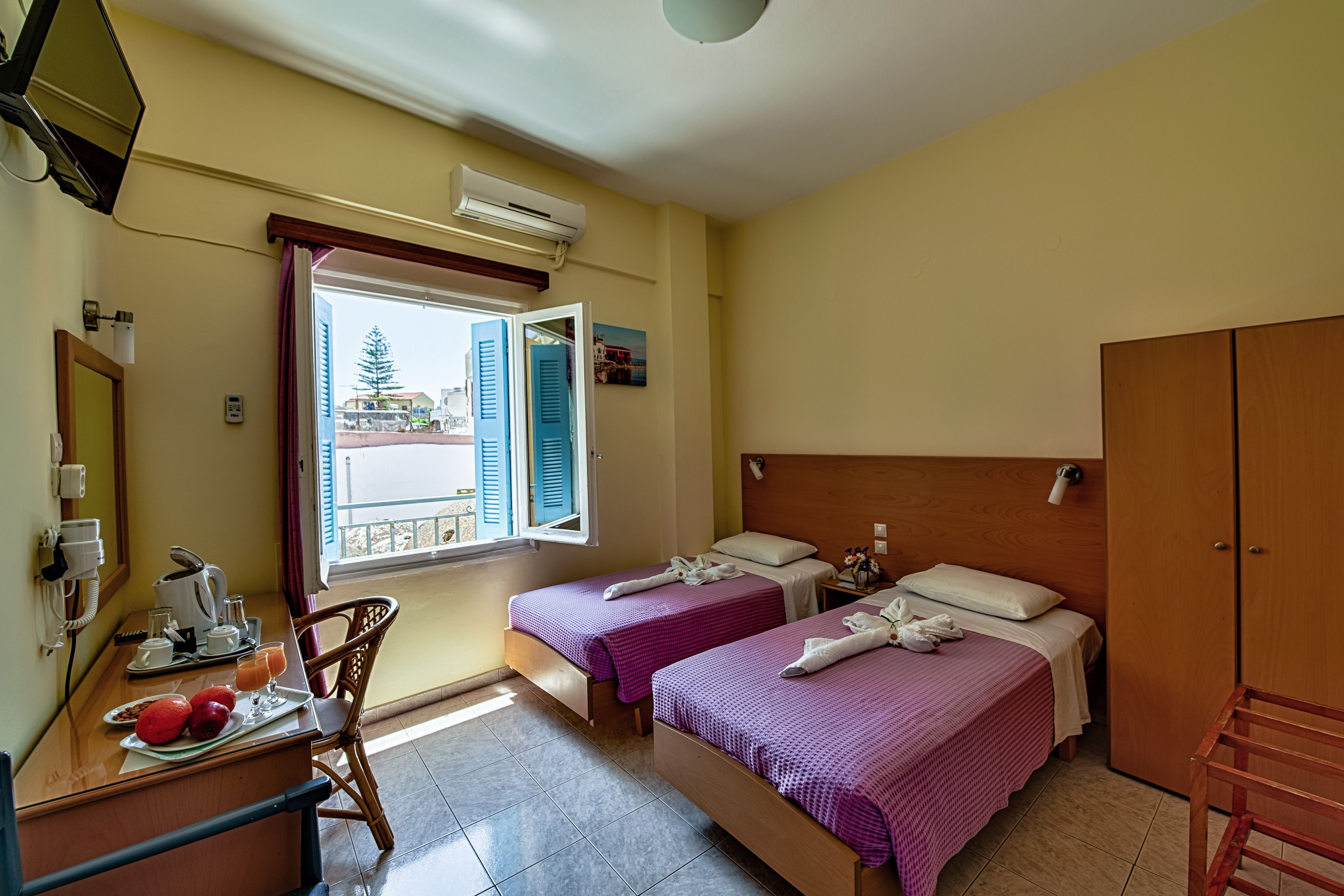 A hotel room with two beds, a desk with breakfast items, a window with shutters, and a wardrobe.