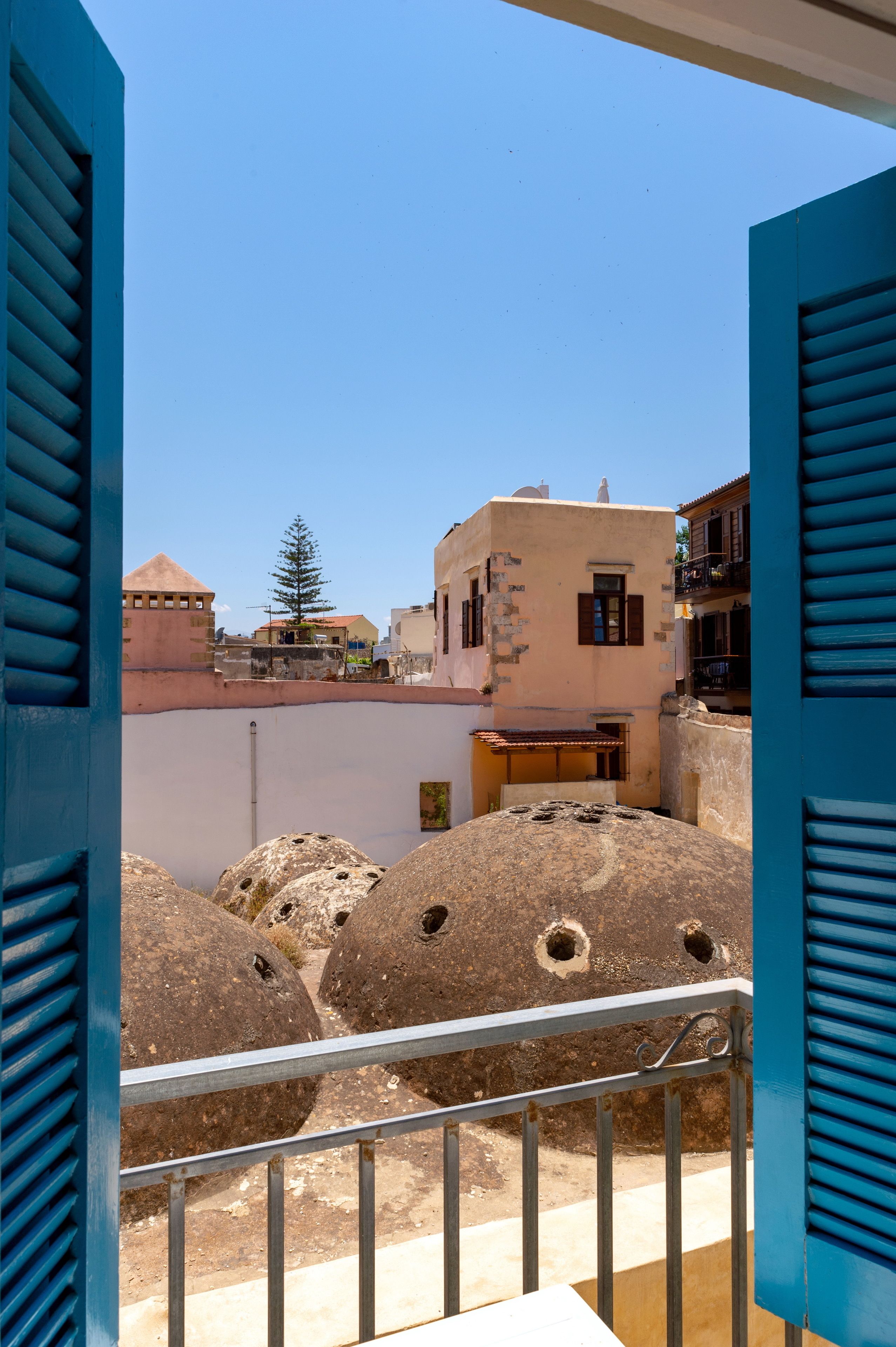 Een balkon met een metalen hekwerk met uitzicht op een erf met grote stenen paaltjes en gebouwen met traditionele architectuur.