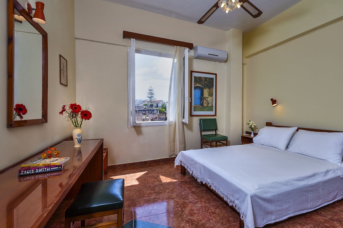 A hotel room with a bed, a desk with a chair, a window with a view, and a painting on the wall.
