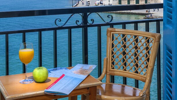 Un balcón con vistas a un pueblo costero, una mesa con un vaso de zumo de naranja, una manzana y un menú.