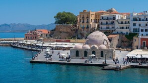 Un paseo marítimo con un edificio histórico que presenta estructuras abovedadas, junto a un mar de aguas azules cristalinas y un telón de fondo montañoso.