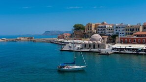 Un pueblo costero con un mar azul cristalino, un velero blanco amarrado en un muelle y edificios históricos de arquitectura abovedada.