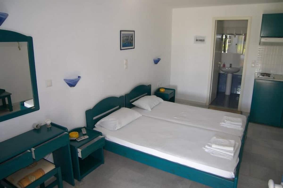 Une chambre d’hôtel avec un lit double, une table de chevet avec une lampe, un petit bureau et une salle de bain visible à travers une porte ouverte.