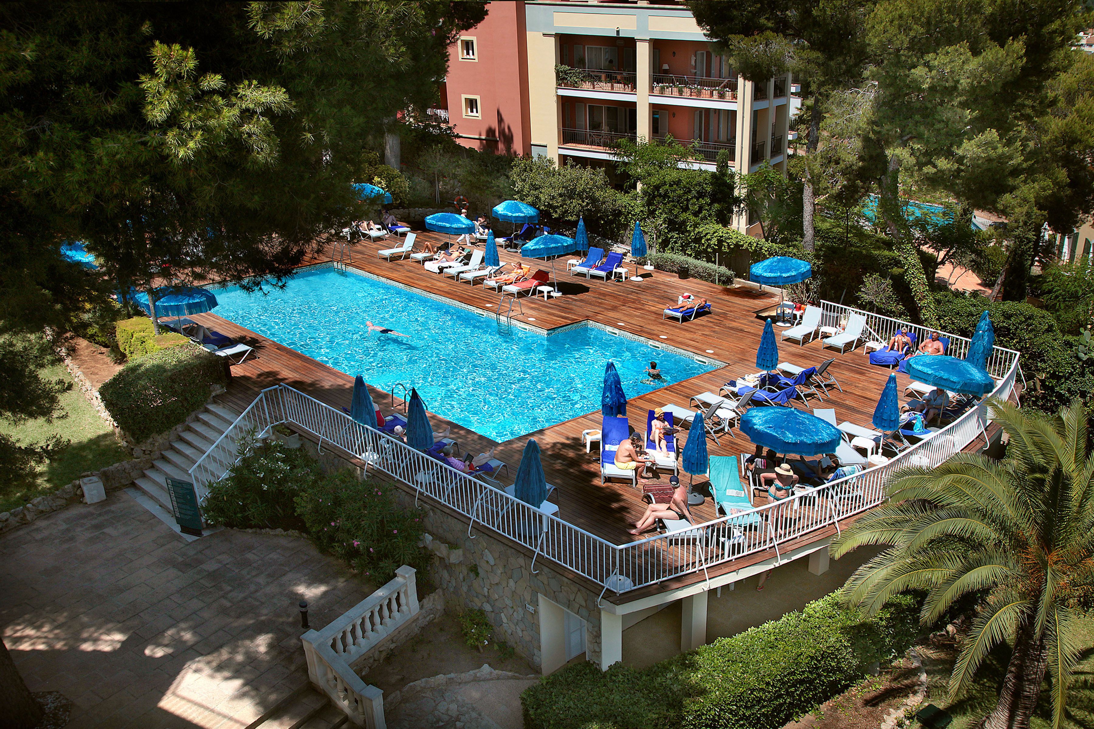 Zona de piscina con sillones de descanso y sombrillas, rodeada de árboles y un edificio al fondo.
