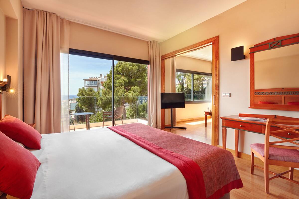 A hotel room with a large bed, a desk, a chair, a TV, and a balcony with a view of buildings and trees.
