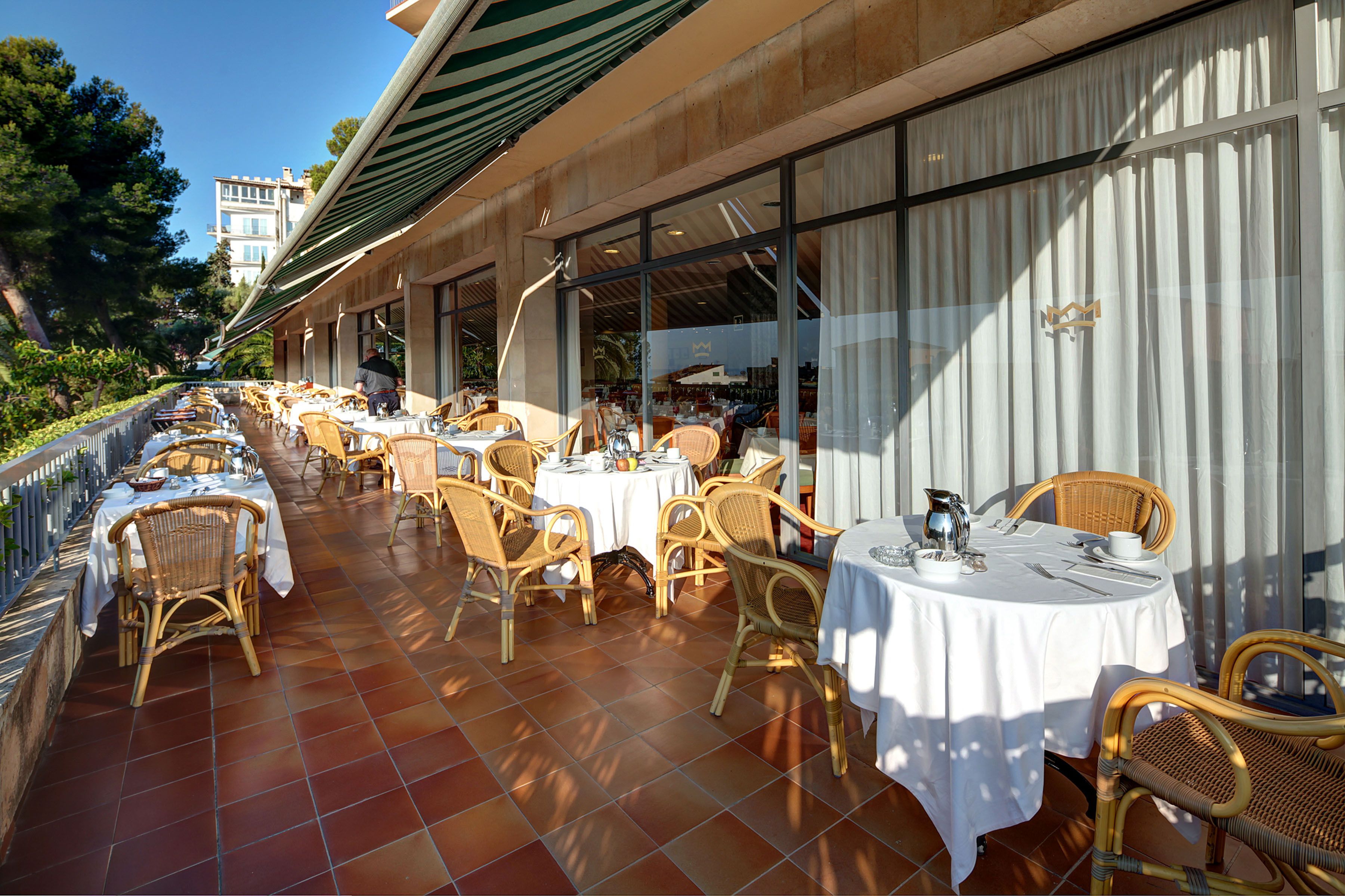 Un espacio al aire libre para comer con sillas y mesas de mimbre, piso de baldosas y vista a un edificio en el fondo.