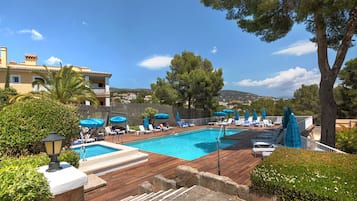 A swimming pool with lounge chairs and umbrellas, surrounded by greenery and a building with a tiled roof.