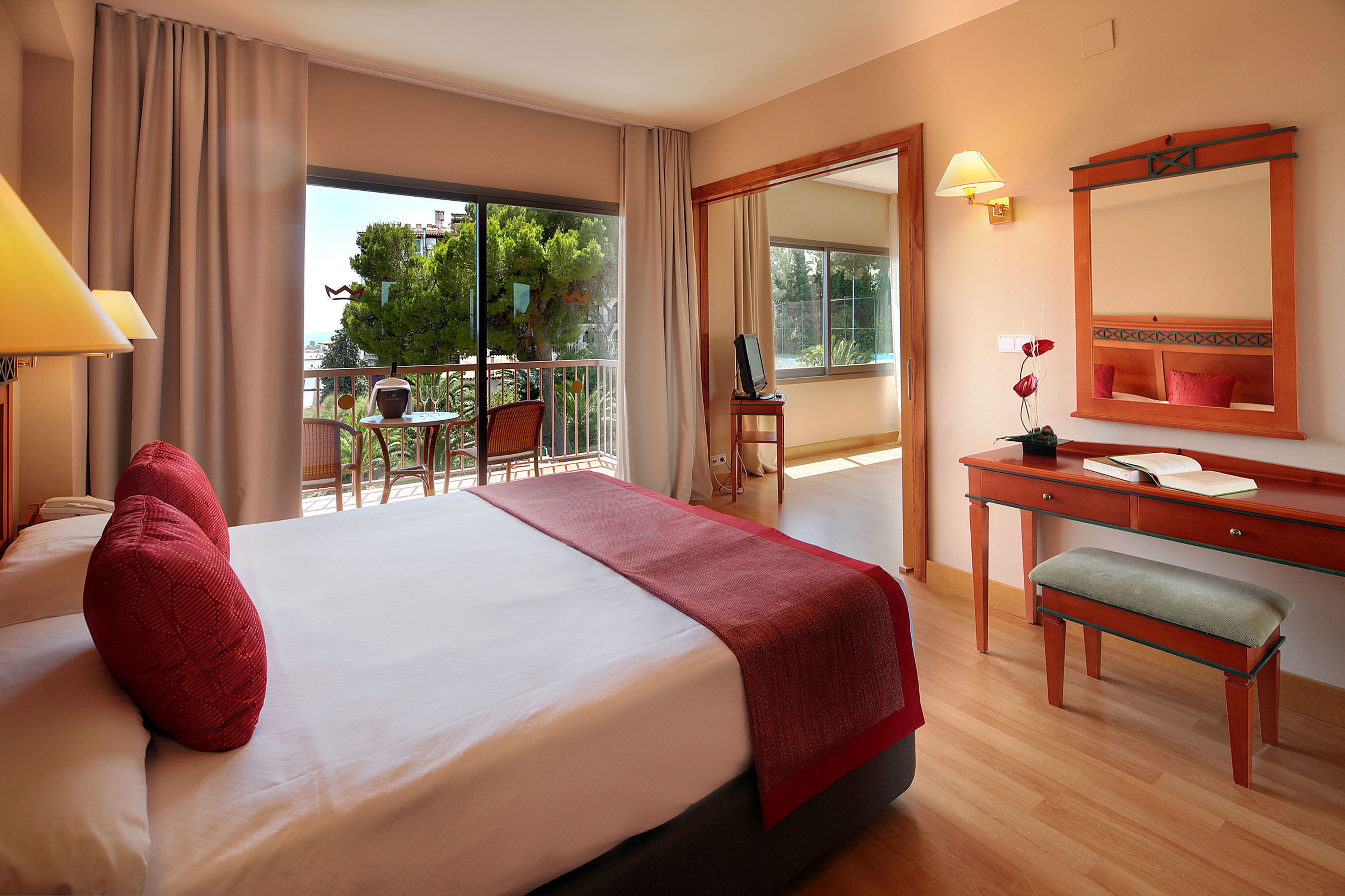 A hotel room with a large bed, a desk with a chair, a balcony with a table and chairs, and a view of trees and buildings.
