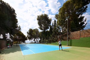 Una cancha de tenis al aire libre con red, rodeada de árboles y un muro de piedra.