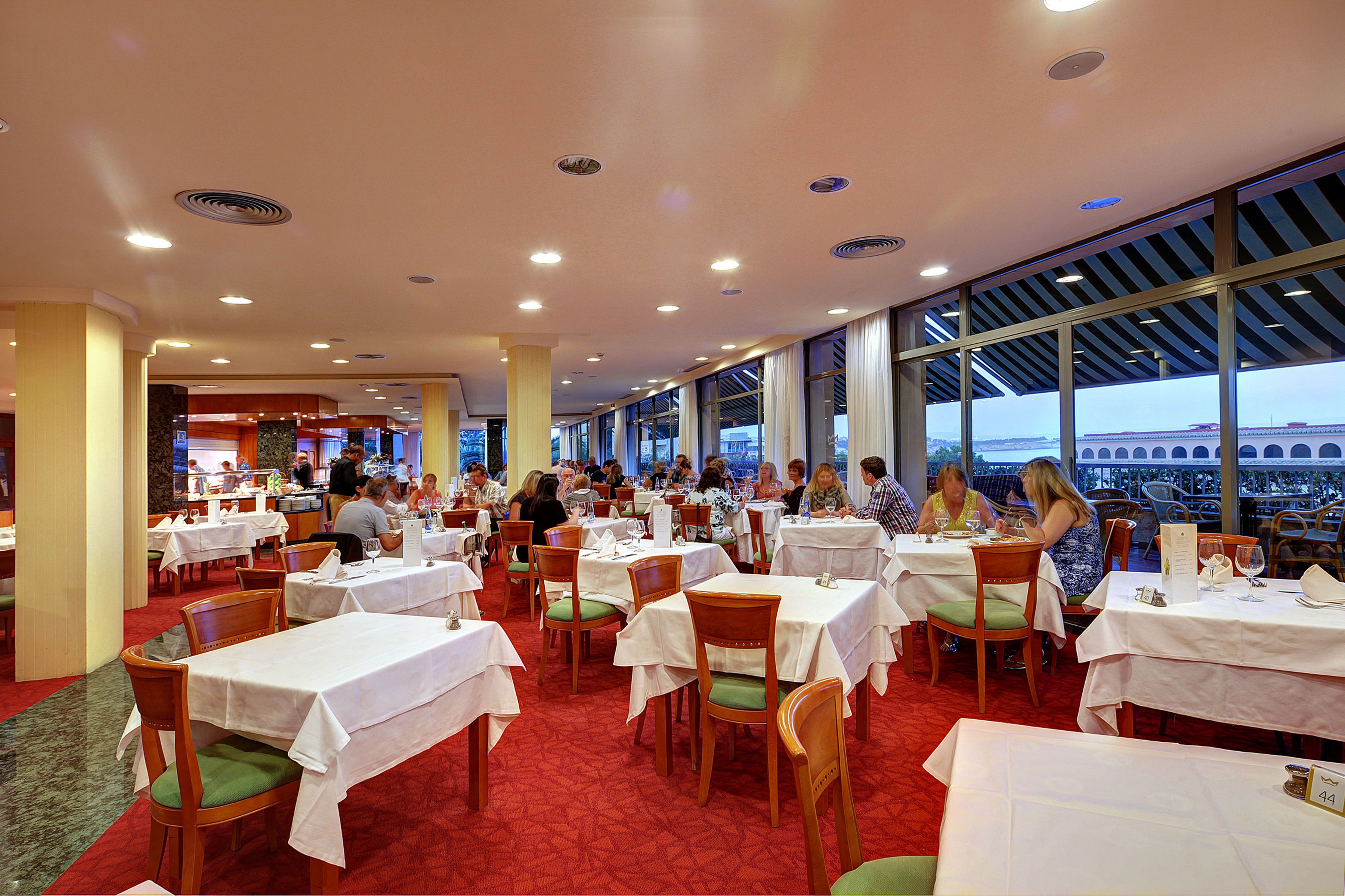 A spacious restaurant with multiple tables and chairs, large windows offering views, and a red carpeted floor.