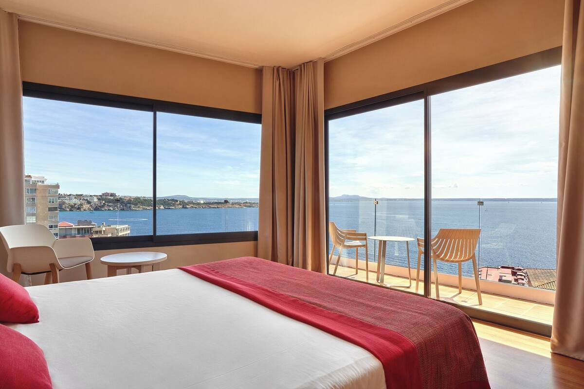 A hotel room with a large bed, a balcony with a table and chairs, and a view of the sea and buildings.