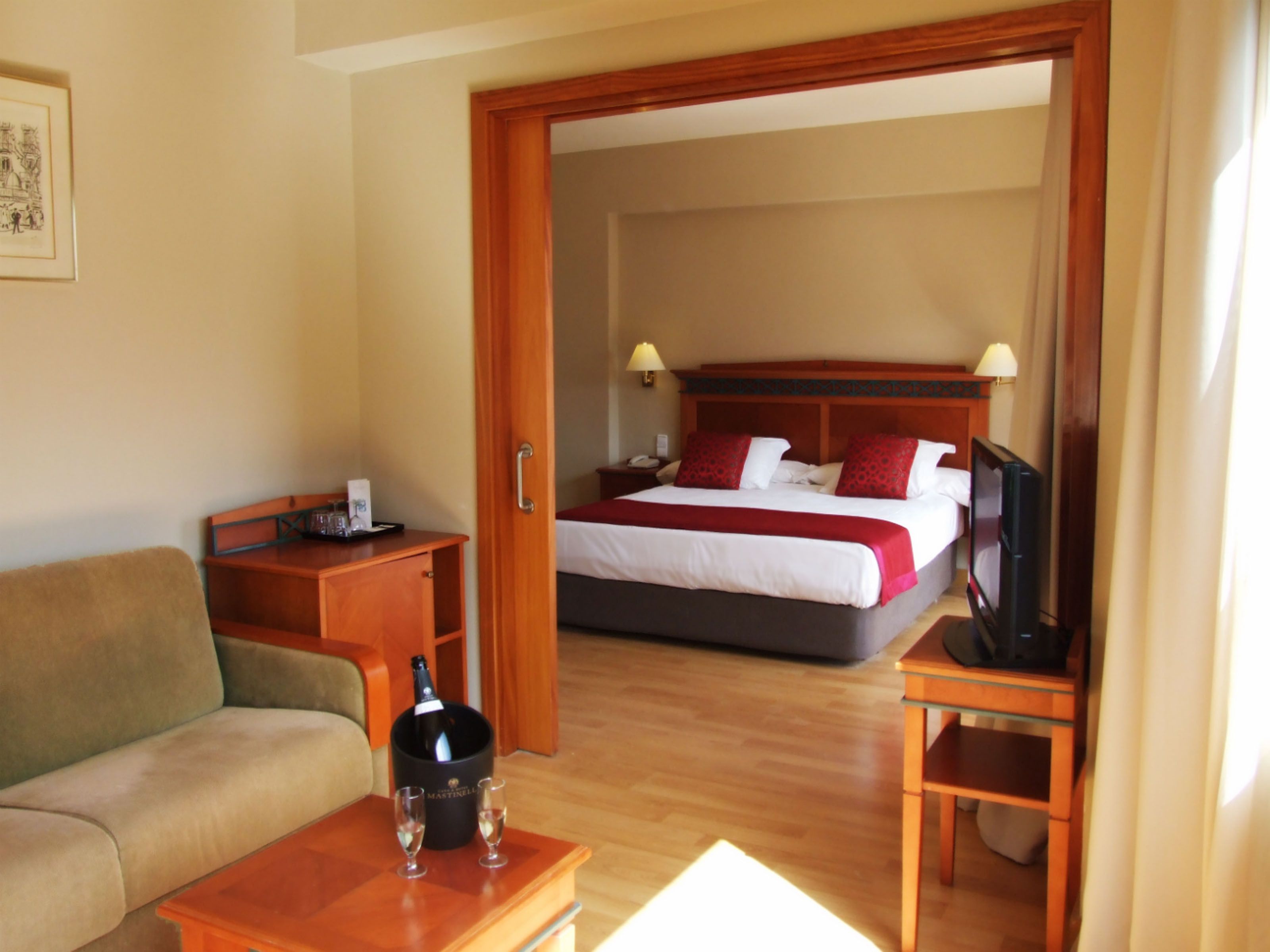 A hotel room with a bed, a TV, a desk with a bottle of champagne, and a sofa.