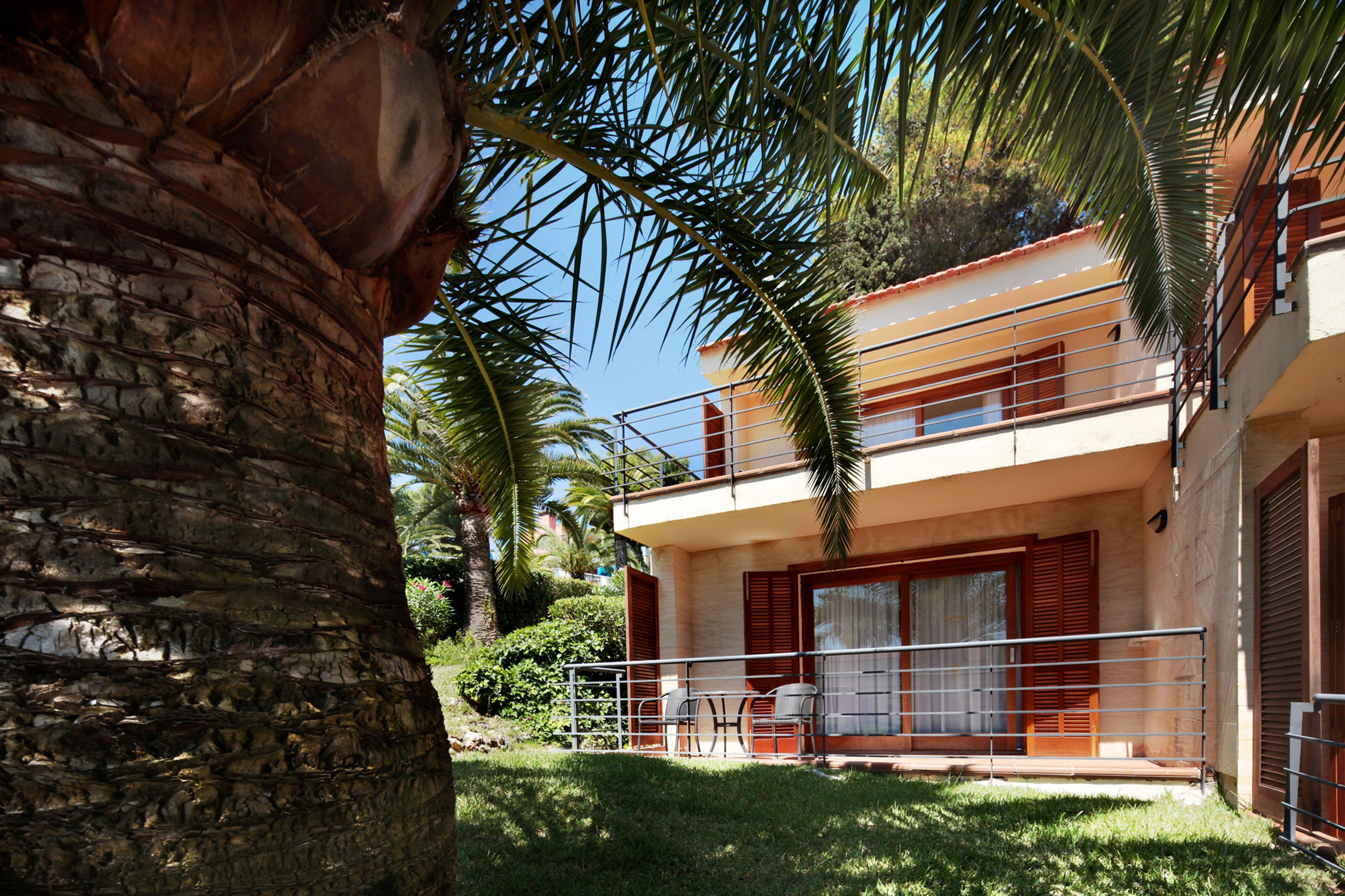 Edifici de dues plantes amb un balcó, una petita zona de pati amb taula i cadires, i una palmera al primer pla.