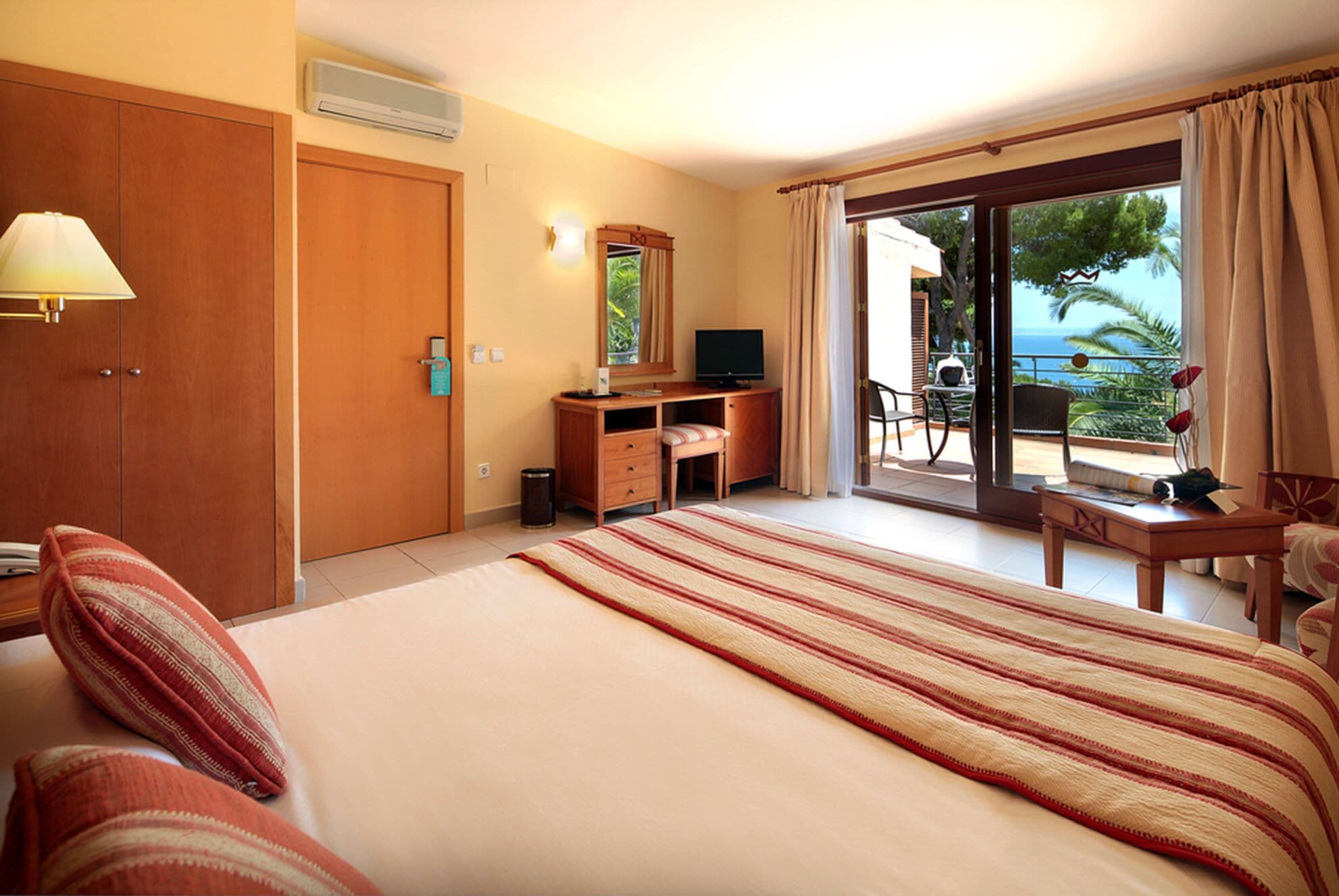 Habitación de hotel con una cama grande, un escritorio con televisor, una ventana con vista al océano y un balcón con mobiliario exterior.