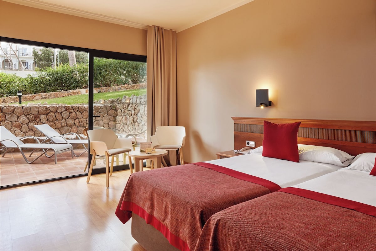 A hotel room with two beds, a large window with a view of the outdoors, a stone wall, and a seating area with chairs and a table.