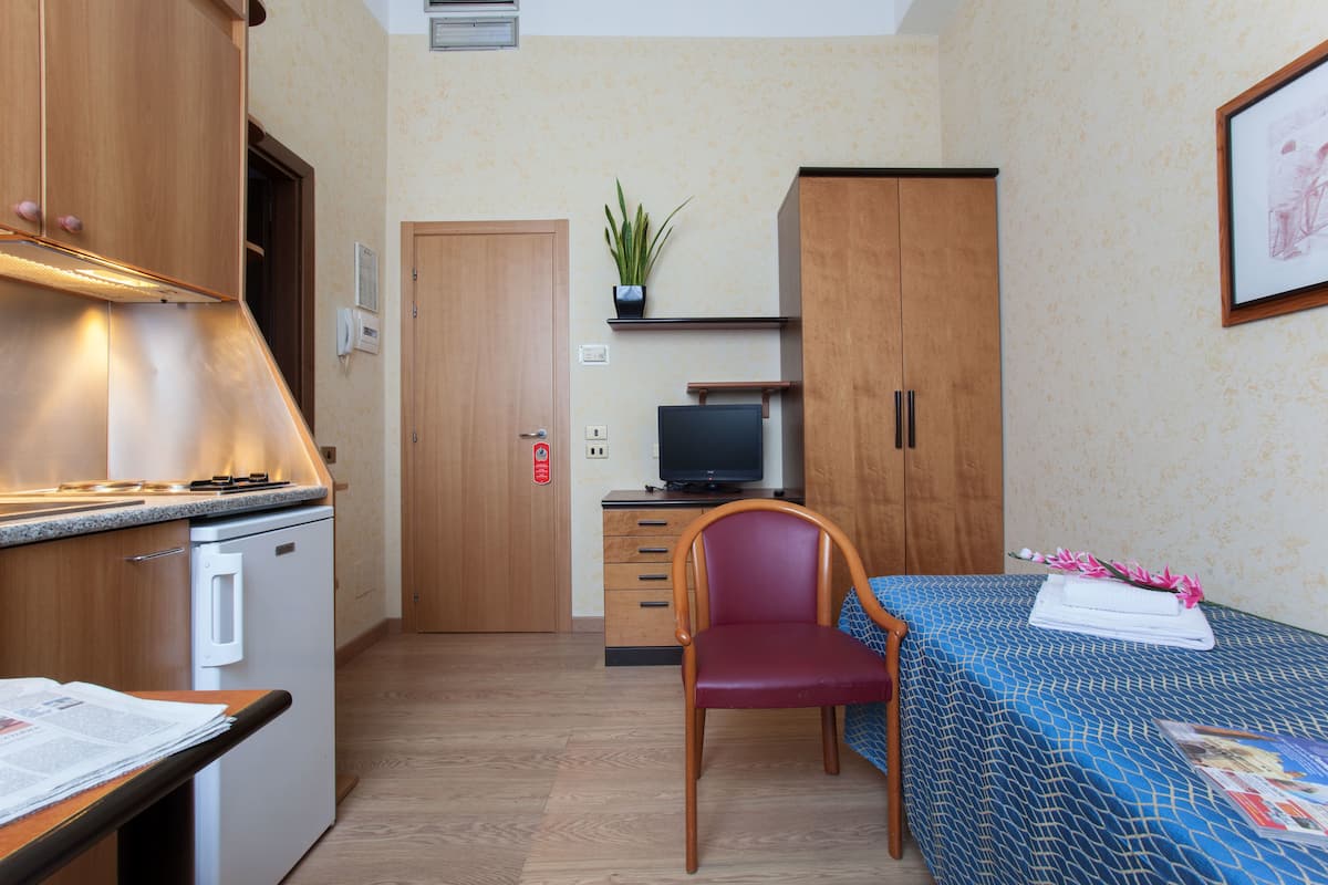 A compact room with a kitchenette, a single bed, a desk with a chair, and a television.