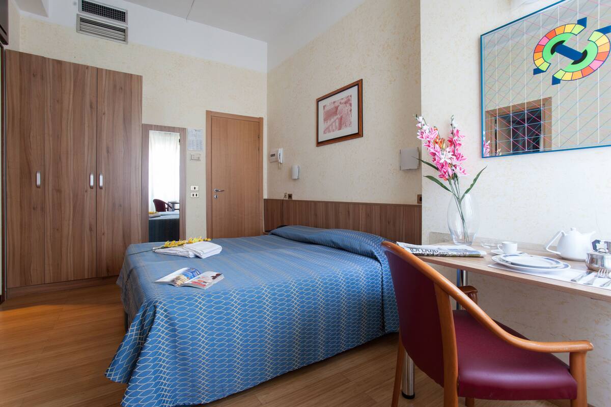 A hotel room with a bed, a desk with a chair, a mirror, and a vase with flowers.
