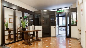 A lobby with a polished tile floor, a wooden reception desk, potted plants, and a glass door leading outside. - Residence Lepontina (Milan)