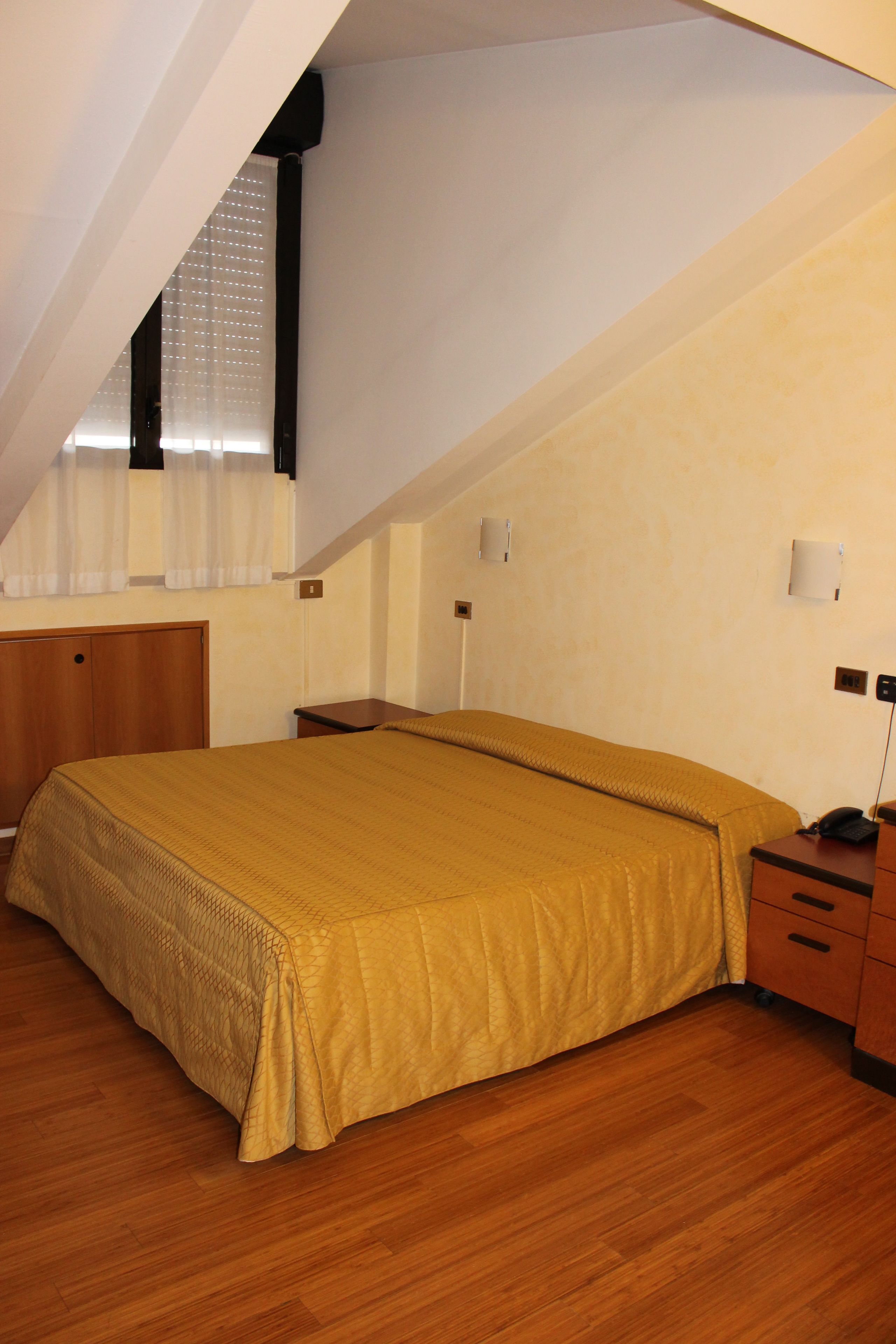 Un dormitorio con una cama grande, suelo de madera, una ventana con cortinas y una mesita de noche con lámpara.