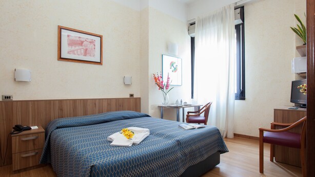 A hotel room with a large bed, a desk with a chair, a TV, and a window with curtains. - Residence Lepontina (Milan)