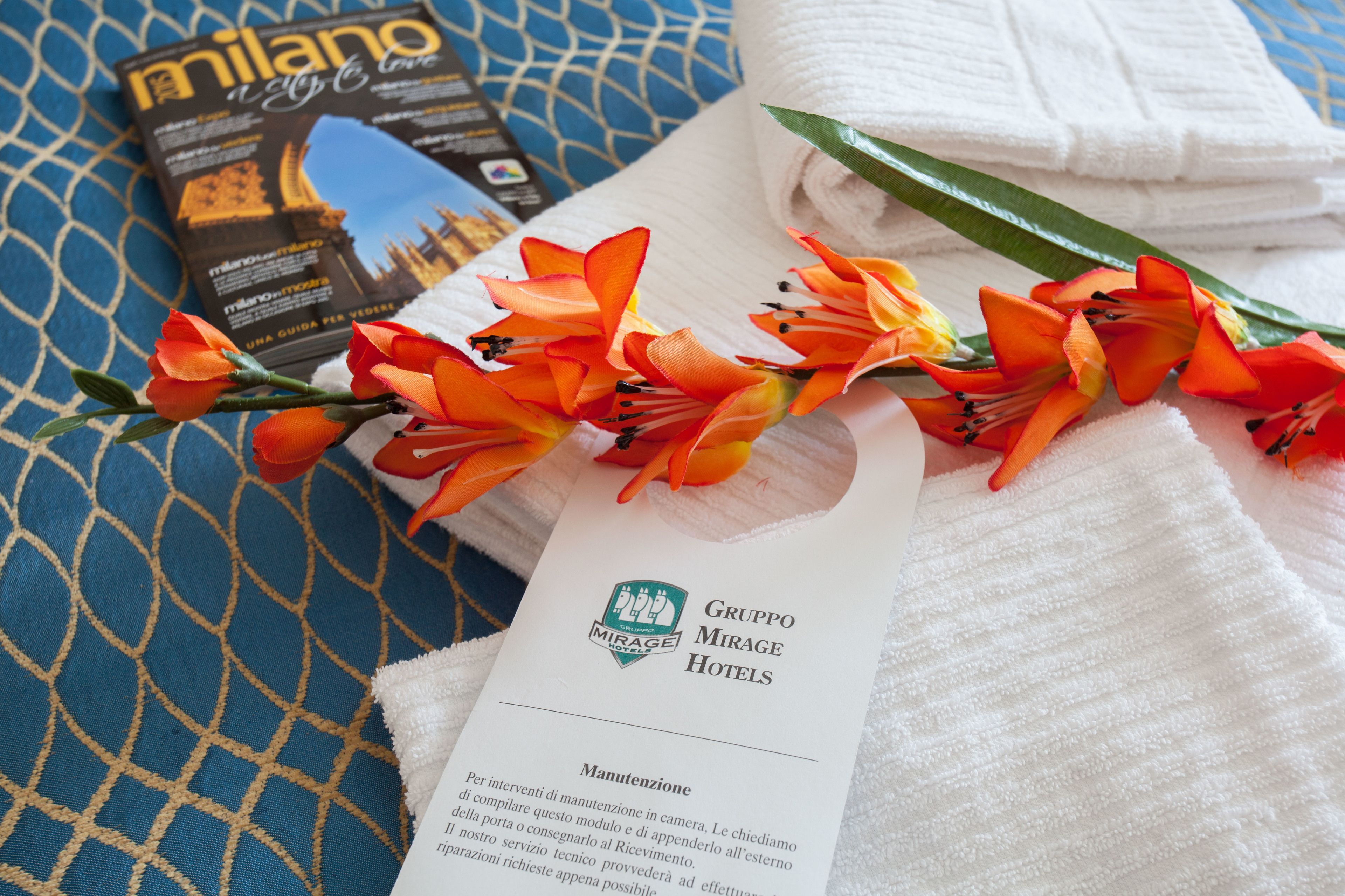 Un folleto de hotel, una toalla doblada y flores naranjas sobre una tela estampada.
