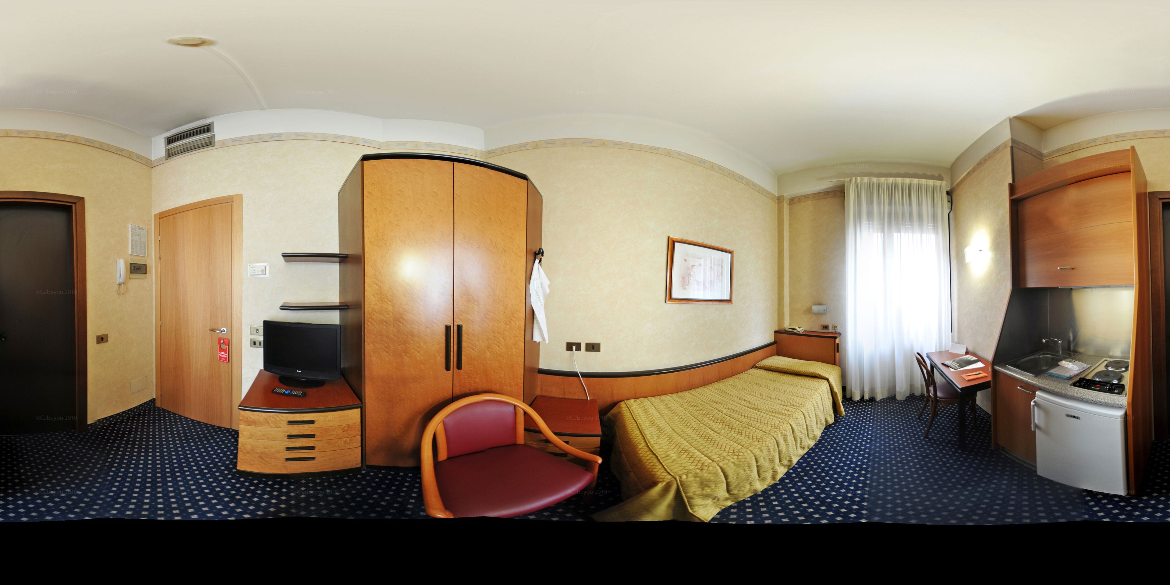 Ein kompaktes Hotelzimmer mit Bett, Schreibtisch, Stuhl, Fernseher, Minikühlschrank und einer Küchenzeile.