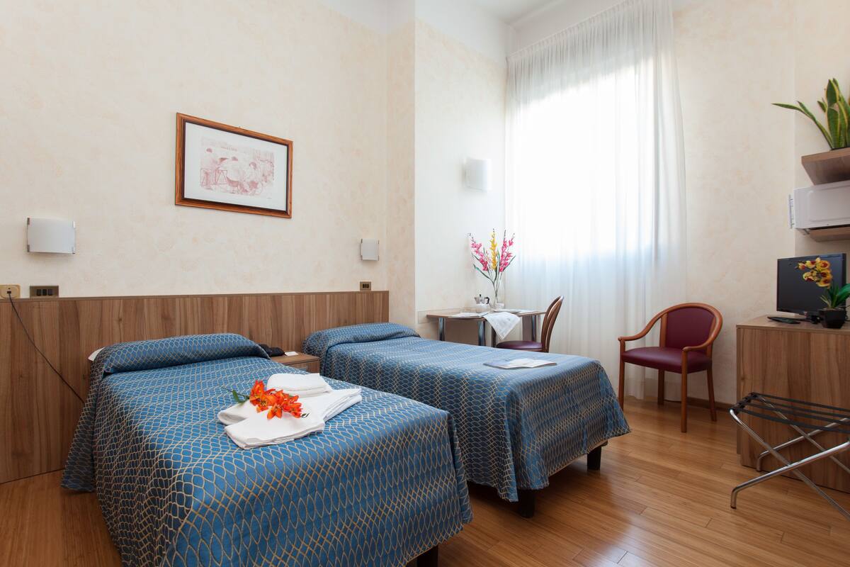 A hotel room with two beds, a desk, a chair, a TV, and a window with curtains.