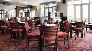 A restaurant with red upholstered chairs and tables, a brick wall, a television, and large windows. - Fox Connaught (London)