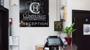 A hotel reception desk with a sign reading "Connaught House Hotel Reception," a security camera, a potted plant, a cash register, and a phone. - Fox Connaught (London)