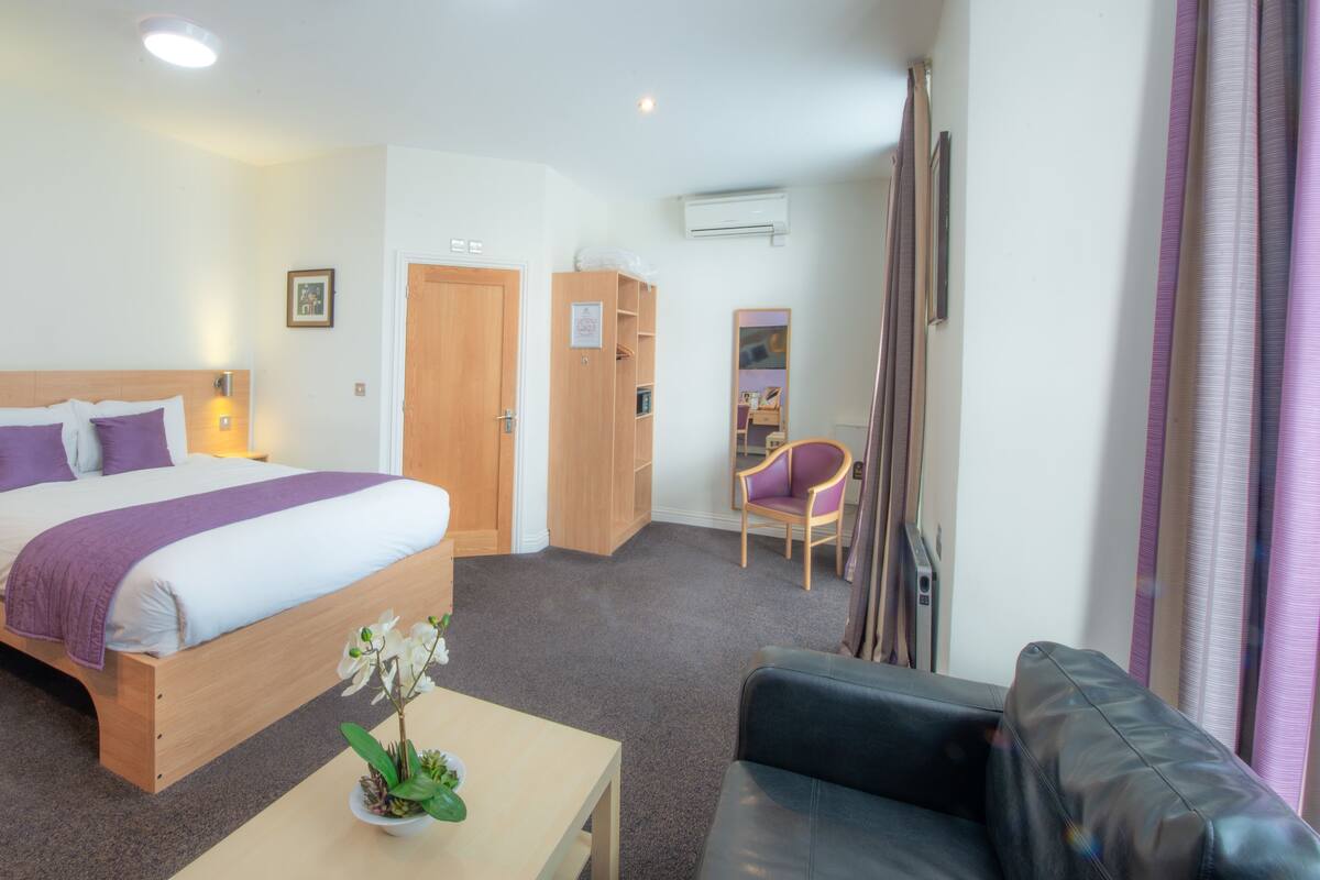 A hotel room with a bed, bedside tables, a desk, a chair, a mirror, and a potted plant.