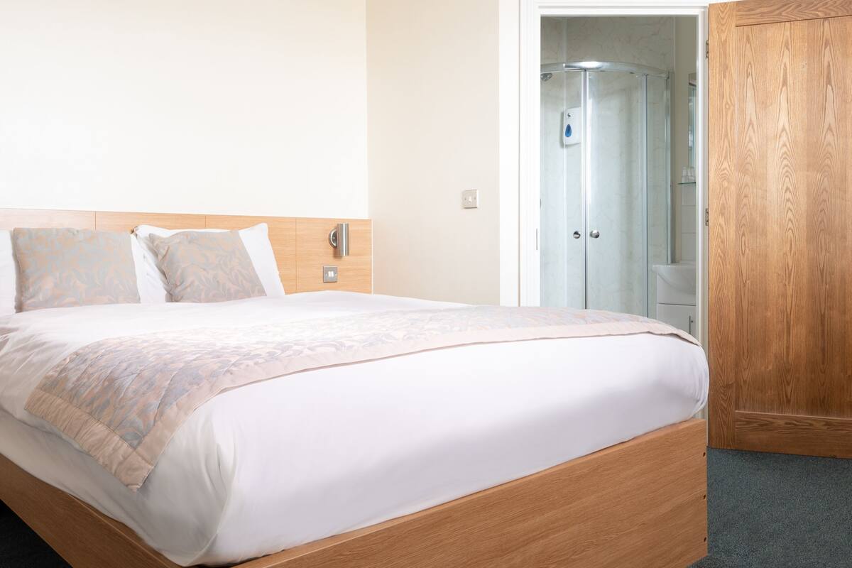 A neatly made bed with a wooden headboard, a wooden wardrobe, and a bathroom visible through an open door.