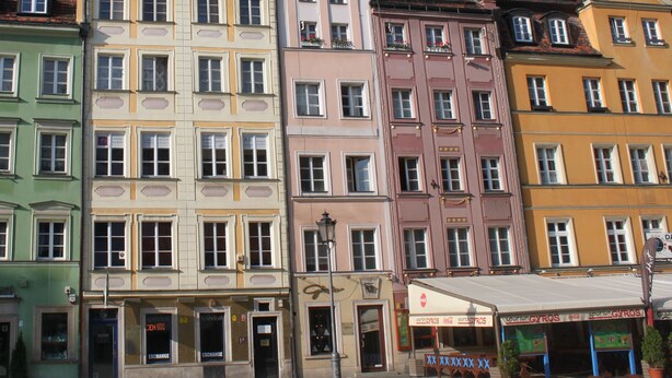 A row of colorful, multi-story buildings with various storefronts, including a cafe and a gym. - Apartment Central (Wroclaw)