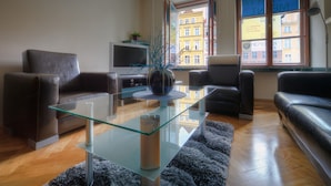 A modern living room with a glass coffee table, leather sofas, a flat-screen TV, and a view of a building through a window. - Apartment Central (Wroclaw)