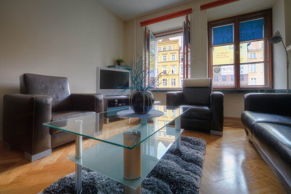 A modern living room with a glass coffee table, leather sofas, a flat-screen TV, and a view of a building through a window.