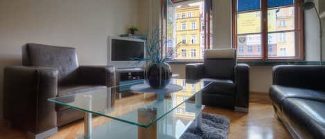 A modern living room with a glass coffee table, leather sofas, a flat-screen TV, and a view of a building through a window.