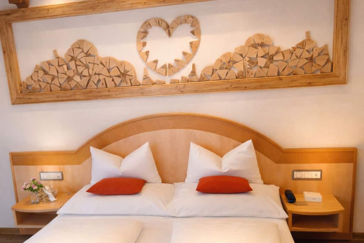 Een tweepersoonsbed met witte lakens en rode kussens, een houten hoofdbord, nachtkastjes met lampen en een kunstwerk boven het bed.