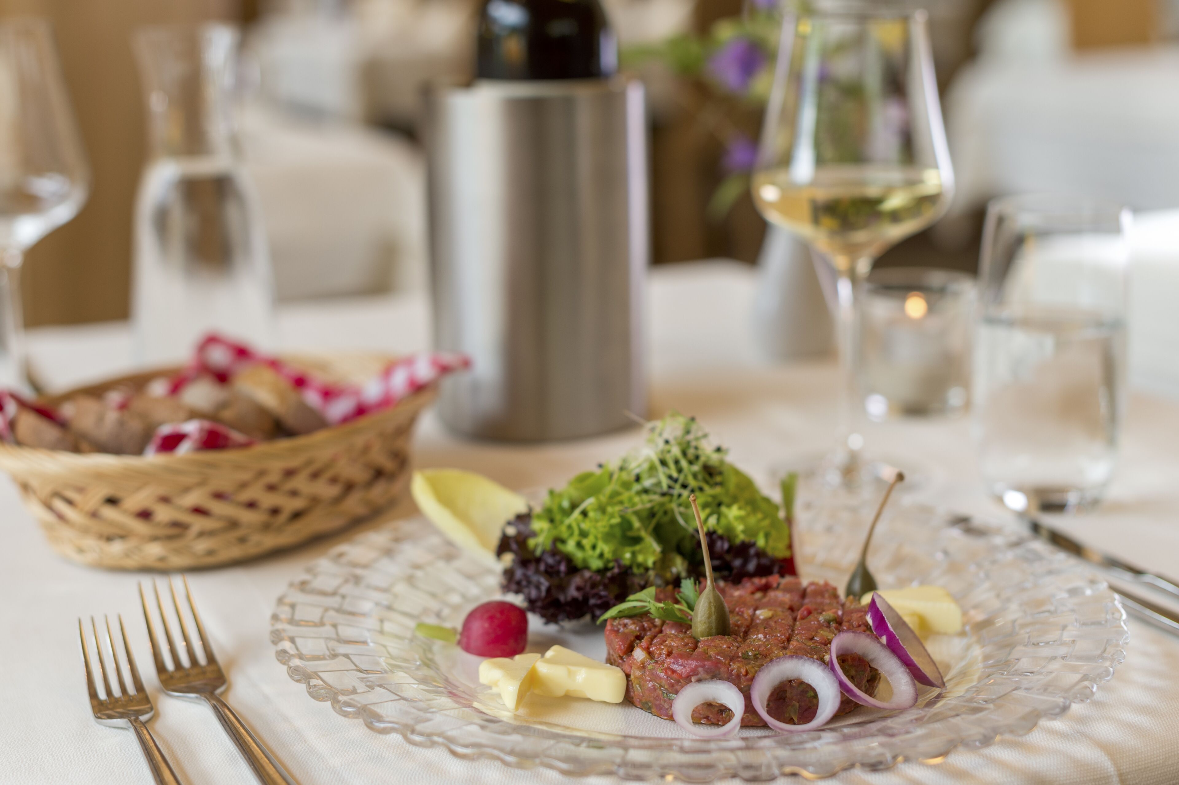 A dining table with a clear glass plate, a basket of bread, and a bottle of wine.