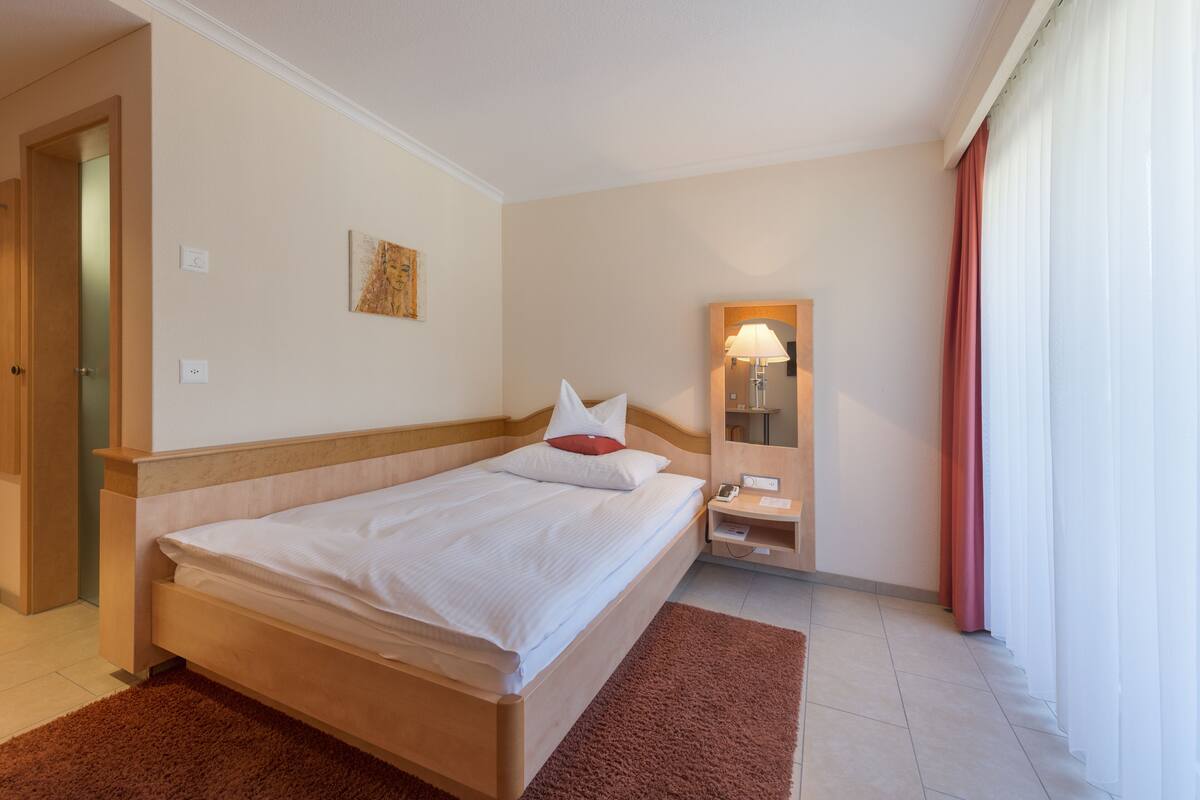 Een hotelkamer met een bed, nachtkastje, spiegel en raam met gordijnen.